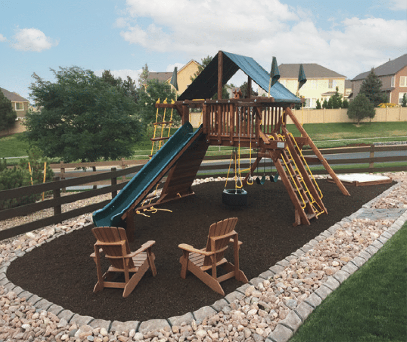 The wooden playground set, complete with slide, swings, and climbing ropes on a mulch surface surrounded by rocks and decorative concrete curbing, sits in a suburban Minnesota neighborhood. Two wooden chairs face the charming play area.