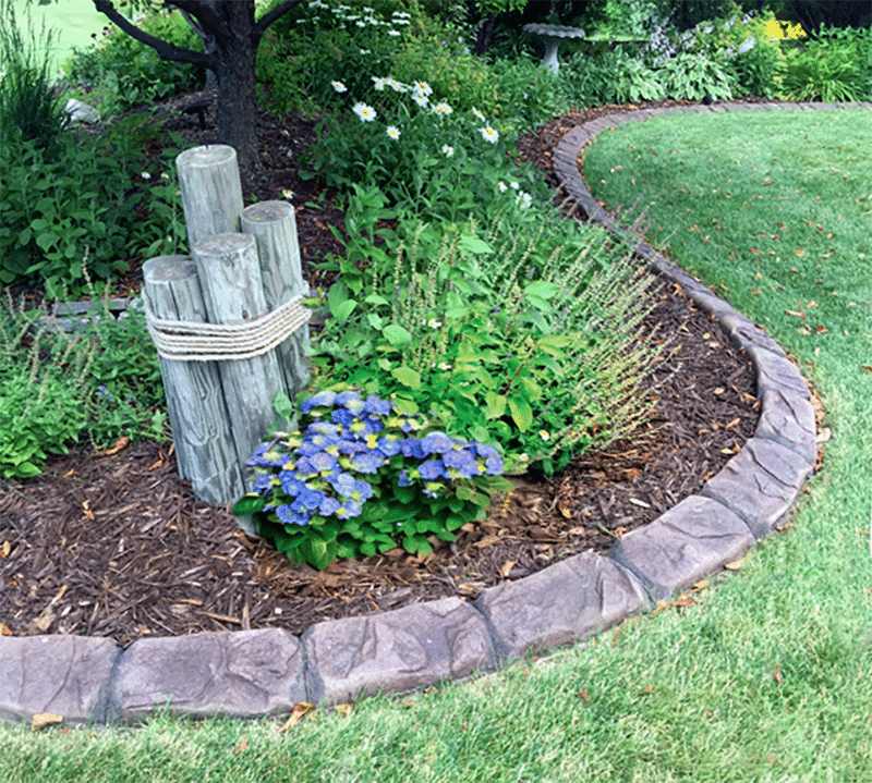 A garden bed featuring vibrant purple flowers is beautifully accented by wooden posts tied with rope. Surrounded by lush grass, the area is tastefully edged with decorative concrete curbing, creating a charming oasis inspired by Minnesota's natural beauty.